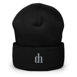 Close-up of a black knit beanie showing its textured fabric and a visible seam, with a partial white logo at the bottom edge.