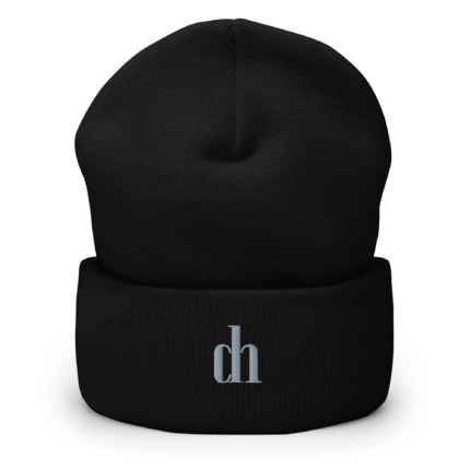 Close-up of a black knit beanie showing its textured fabric and a visible seam, with a partial white logo at the bottom edge.