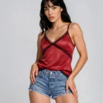 Woman wearing a red satin camisole with black trim and denim shorts, posing against a gray background.