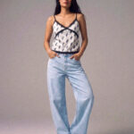 Model wearing a white palm-print camisole with black lace trim and light blue jeans, standing with hands in pockets on a neutral studio backdrop.