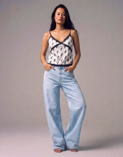Model wearing a white palm-print camisole with black lace trim and light blue jeans, standing with hands in pockets on a neutral studio backdrop.